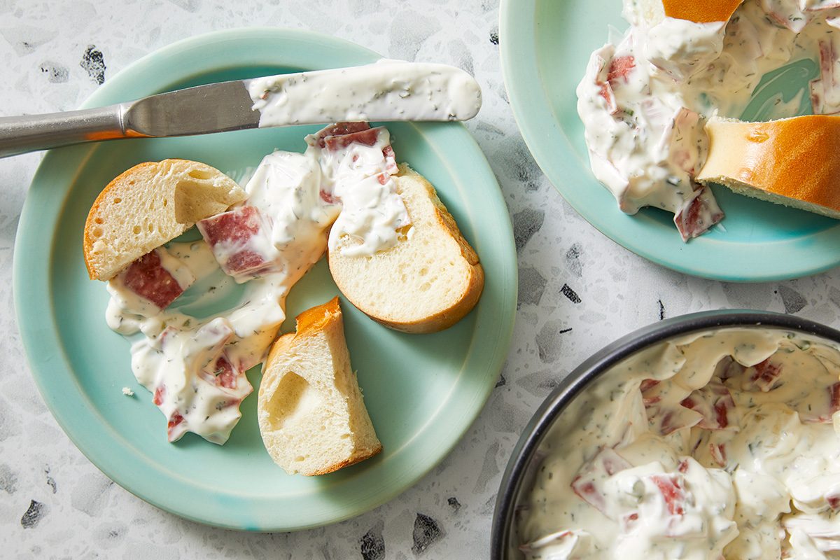 overhead shot of platter of Easy Bagel Dip; surrounded by slices of bagel and bread on a turquoise plate; A knife with a dollop of the dip is placed next to one of the slices, The background is a light, speckled surface
