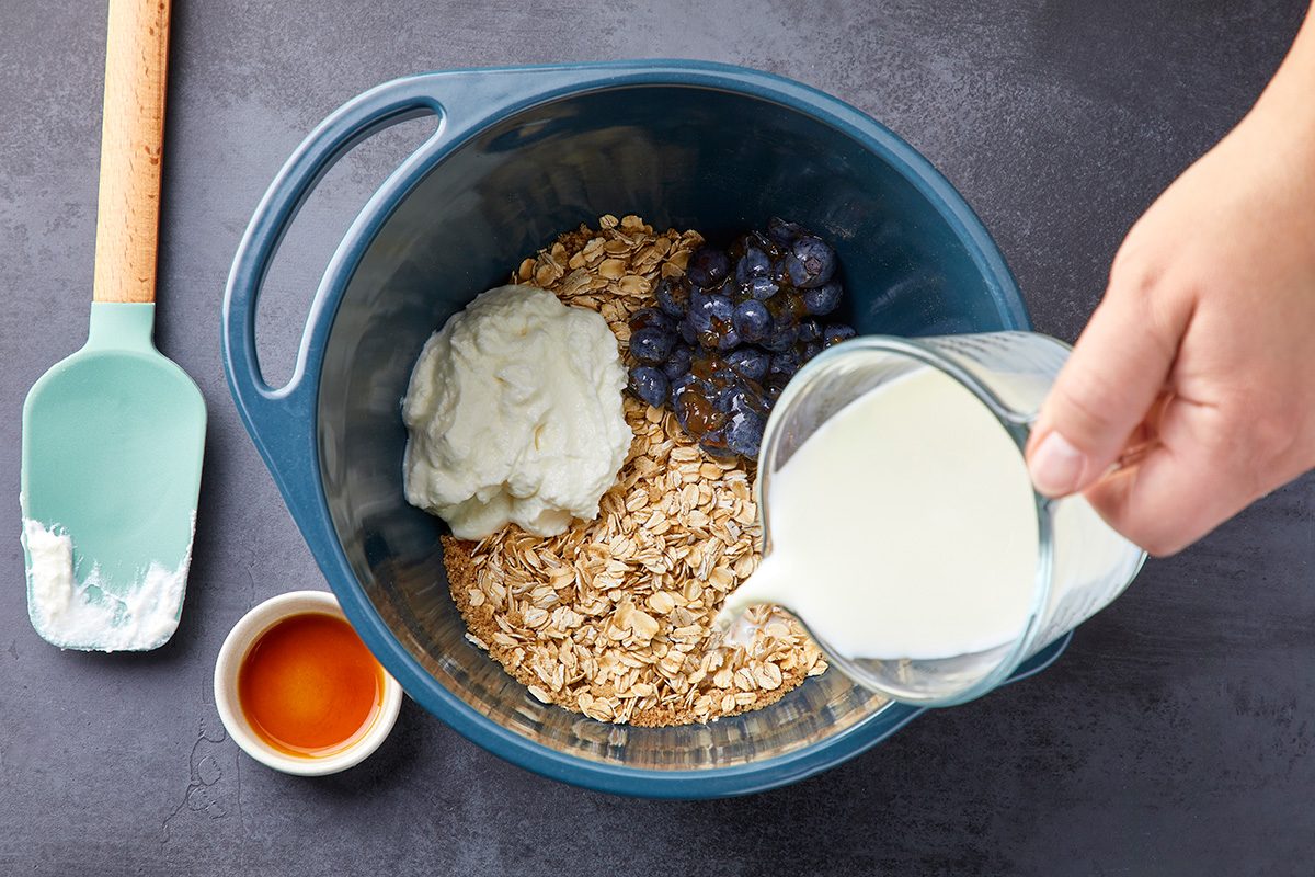 Overhead shot of stir in milk; blueberries; yogurt and vanilla until combined; spatula on left side; all arranged on a dark surface