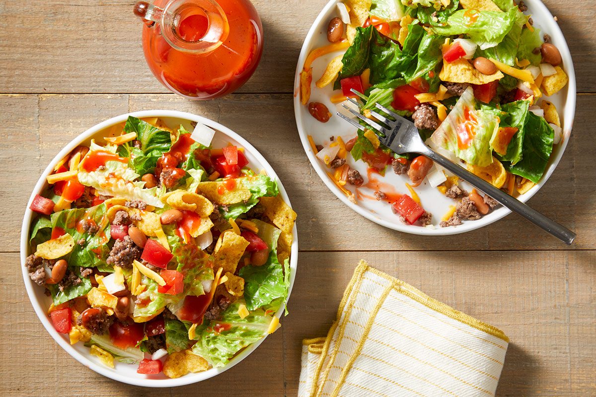 Two plates of taco salad with lettuce, ground beef, cheese, tomatoes, beans, and tortilla chips are on a wooden table next to a jar of dressing and a folded yellow-striped napkin.