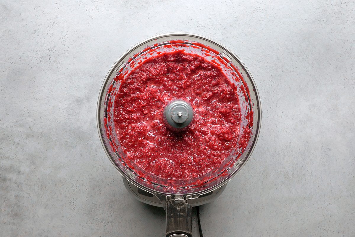 A food processor filled with blended red berry puree sits on a light gray countertop, viewed from above.