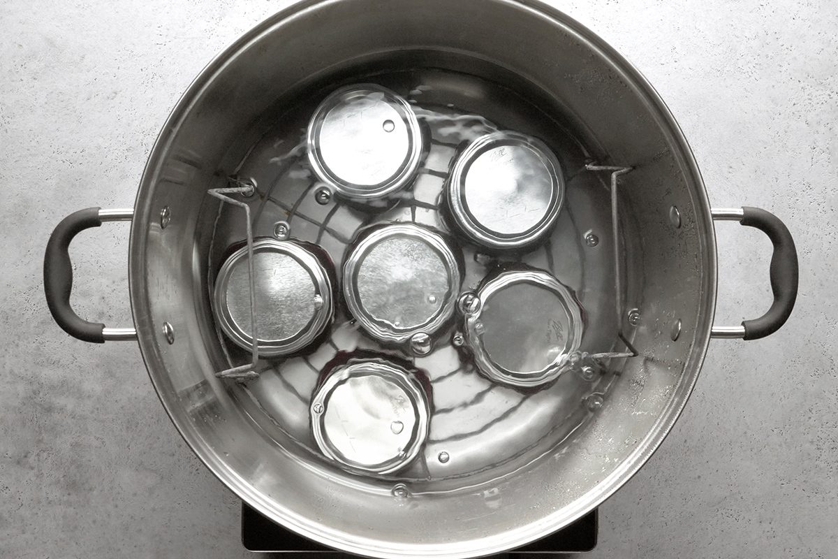 A large metal pot filled with water contains six glass jars with metal lids placed upside down inside on a metal rack, ready for processing or sterilizing.