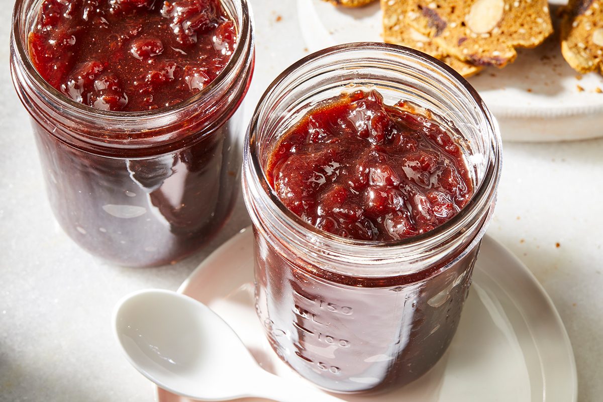 Two glass jars filled with chunky red jam sit on a white plate with a white spoon beside them. In the background, there are crackers with visible nuts.