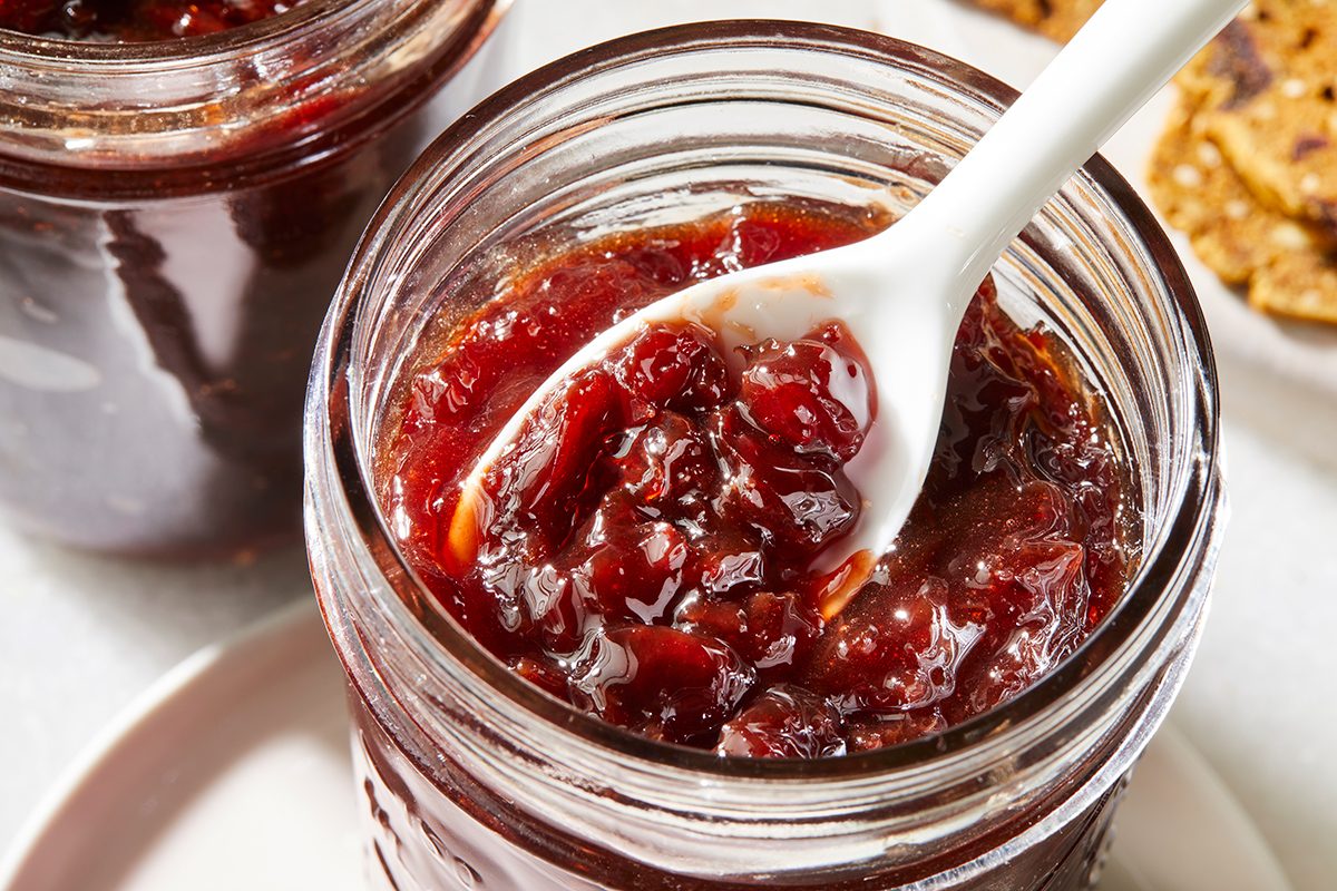 Glass jar filled with chunky red jam or preserves, with a white spoon scooping some out. Another jar and some crackers are partially visible in the background.