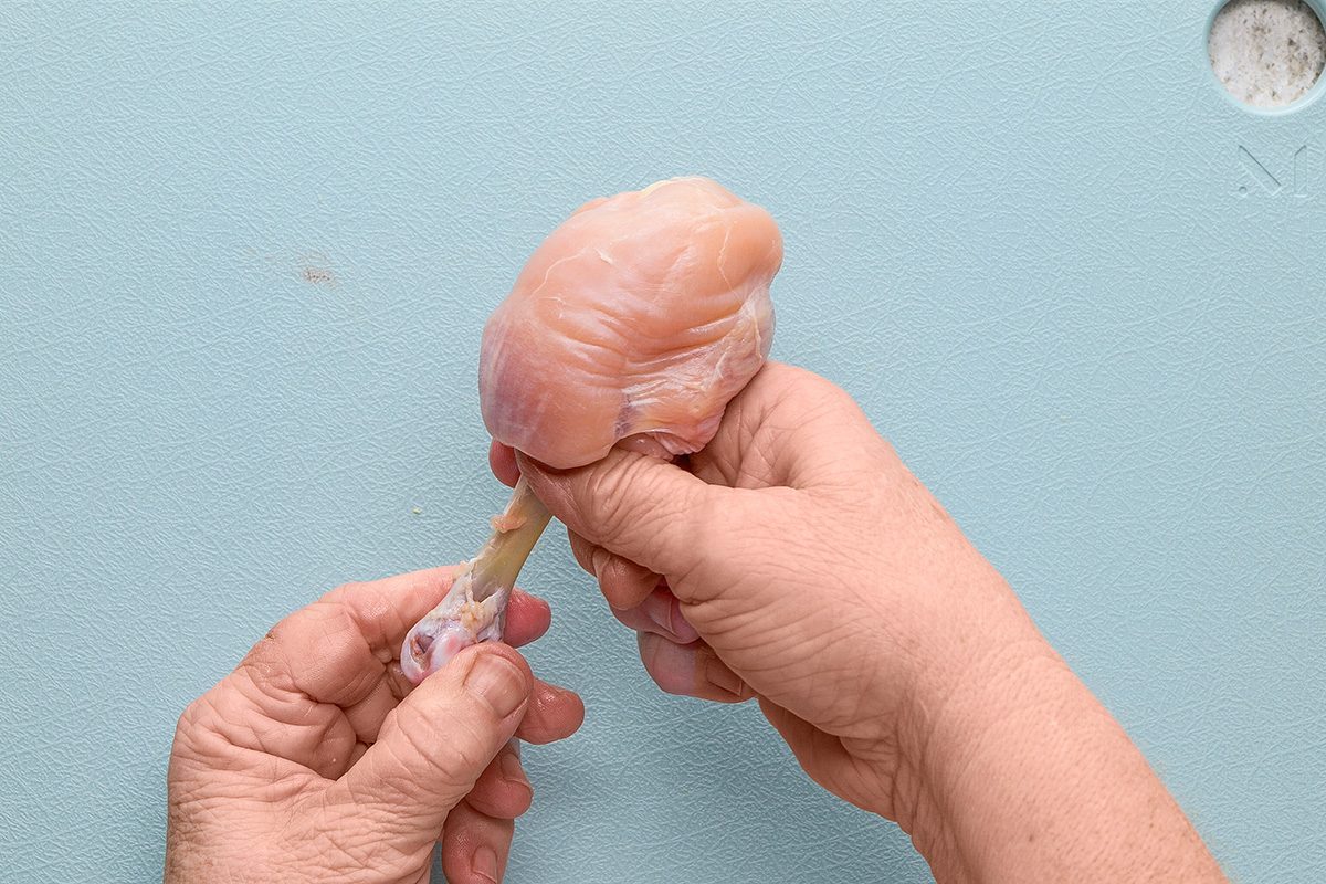 A pair of hands holding a raw chicken drumstick on a light blue cutting board, with one hand gripping the meat and the other holding the bone.