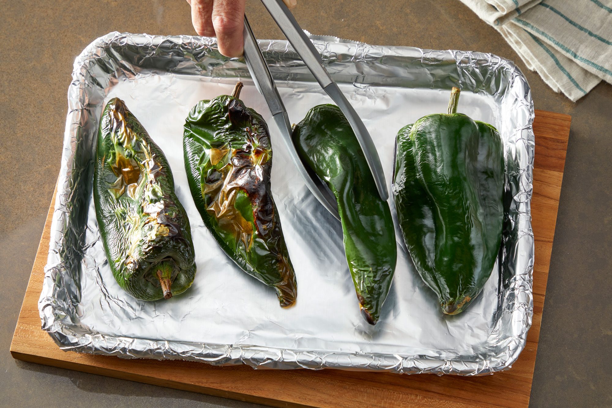 Overhead shot; To make peppers preheat broiler to high; Place poblano peppers on a foil-lined baking sheet; Broil peppers 4 inch from the heat until skins blister 4-5 minutes; With tongs; rotate peppers a quarter turn; Broil and rotate until all sides are blistered and blackened; wooden board; a napkin is nearby; all set on a light brown surface