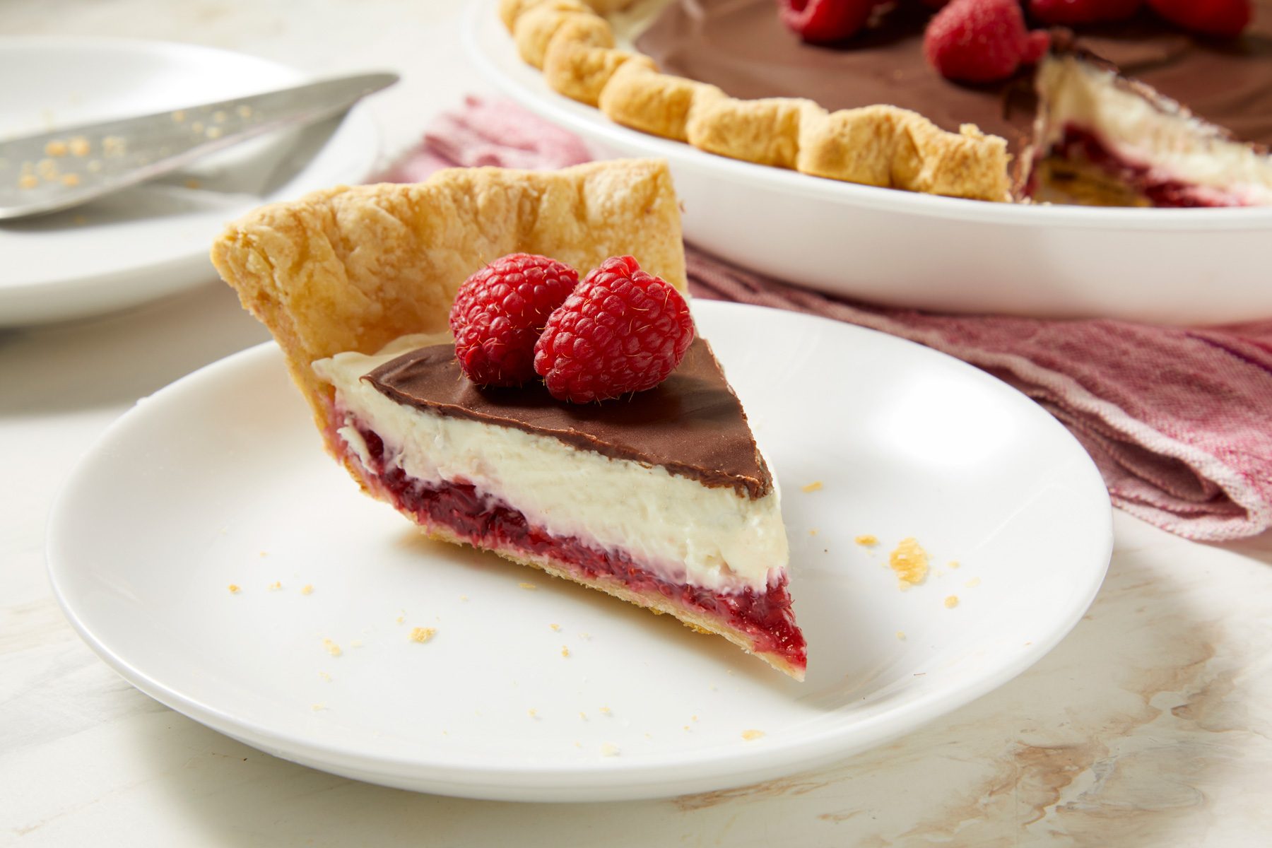 3/4th shot of a slice of Chocolate Raspberry Pie placed on a white plate; garnishing the top of the pie slice are three fresh raspberries, in the background, a whole pie is seen, along with a plate and a fork