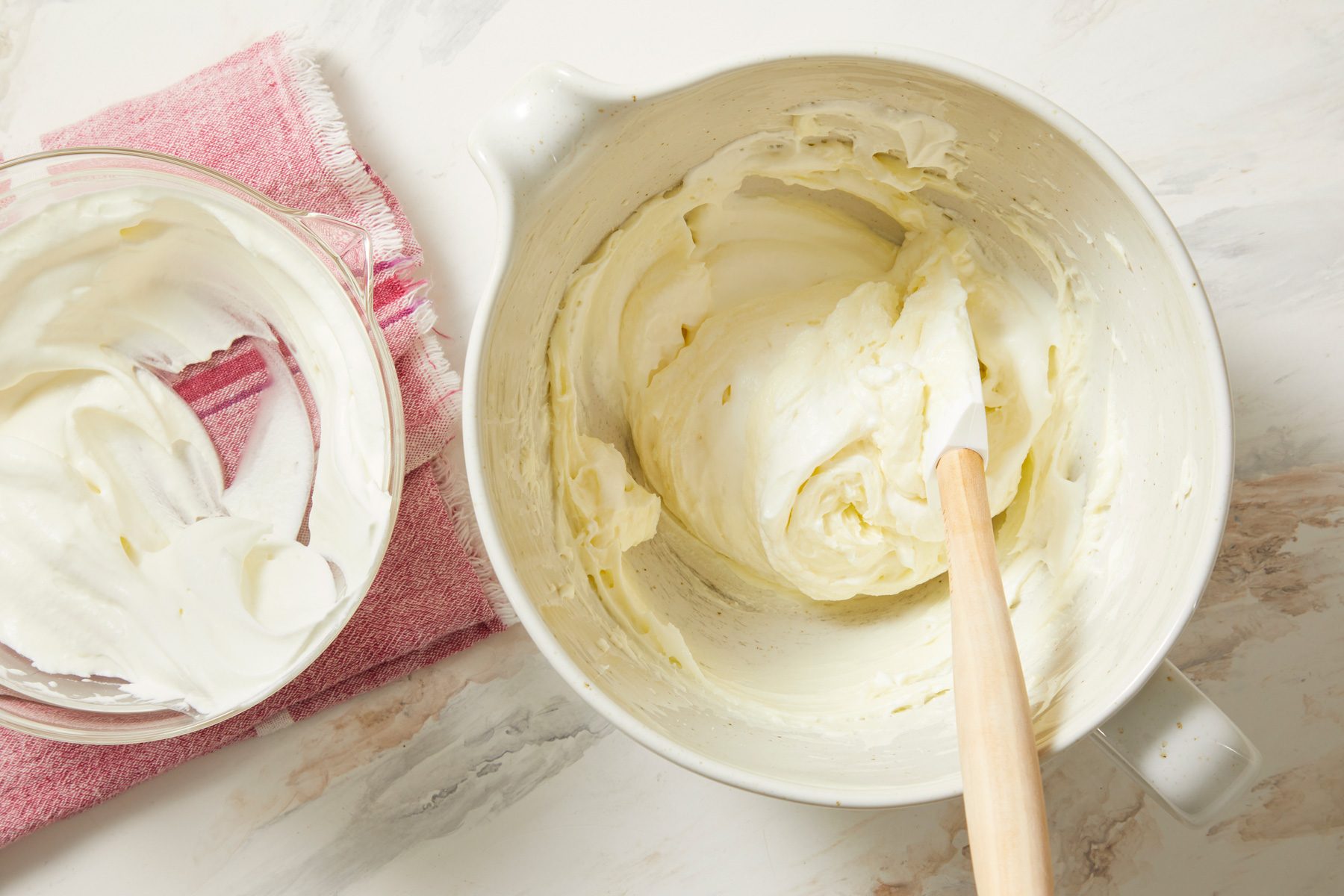 overhead shot of two bowls of creamy mixtures on a light, textured countertop; on the right, a larger bowl contains whipped cream or frosting, with a wooden spatula resting in it; on the left, a smaller glass bowl is holding similar cream, while there is pink dish towel underneath