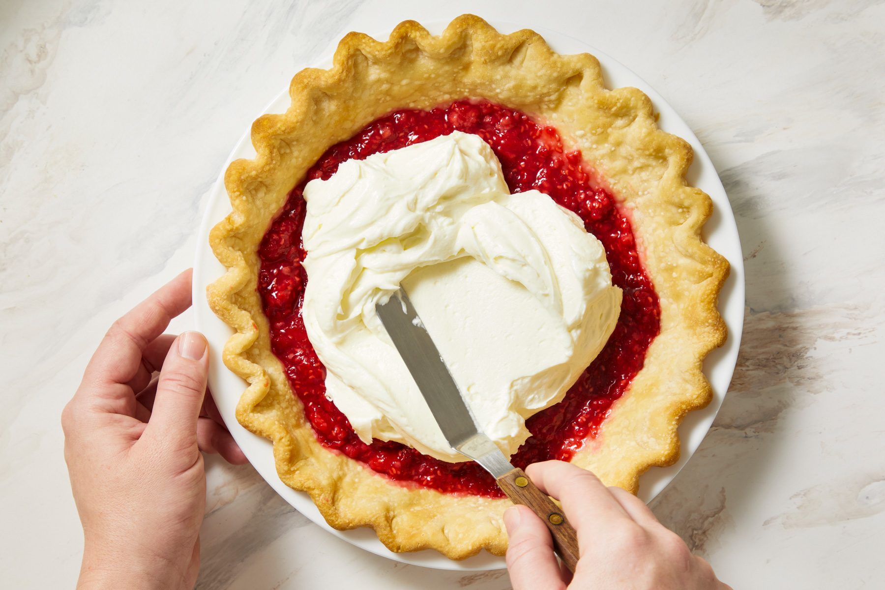 overhead shot of pie sitting in a white dish with a crimped edge; a hand is seen holding a spatula, which is being used to spread the creamy topping over the red filling; the background is a light marbled surface