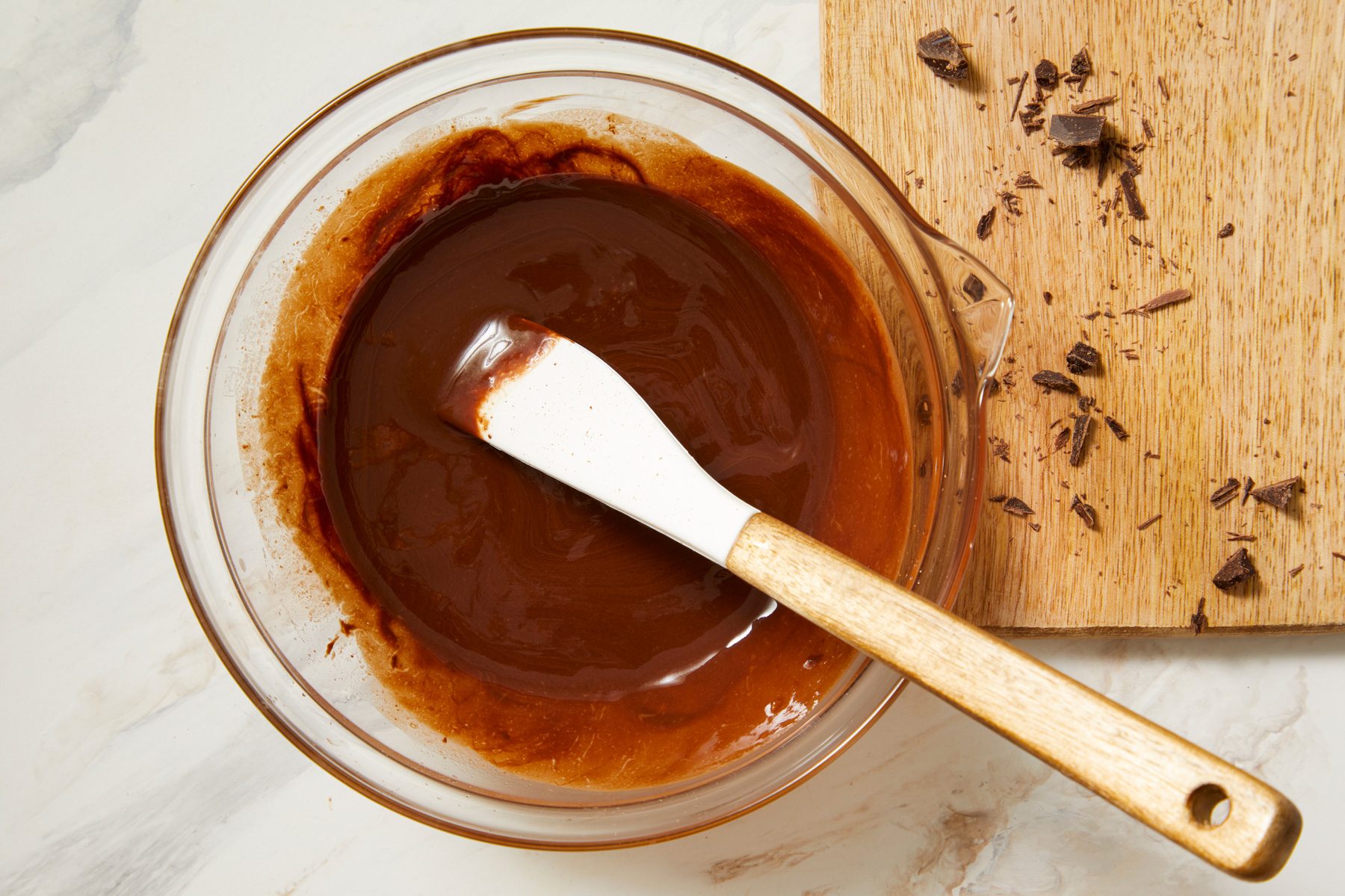 overhead shot of a glass mixing bowl filled with a rich brown chocolate mixture; a white spatula rests inside the bowl, beside the bowl, there is a wooden cutting board scattered with small pieces of chocolate, the background features a light marbled surface