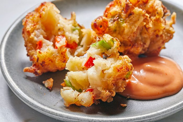 Two golden-brown vegetable fritters, broken open to show colorful filling, sit on a gray plate next to a dollop of creamy orange dipping sauce.
