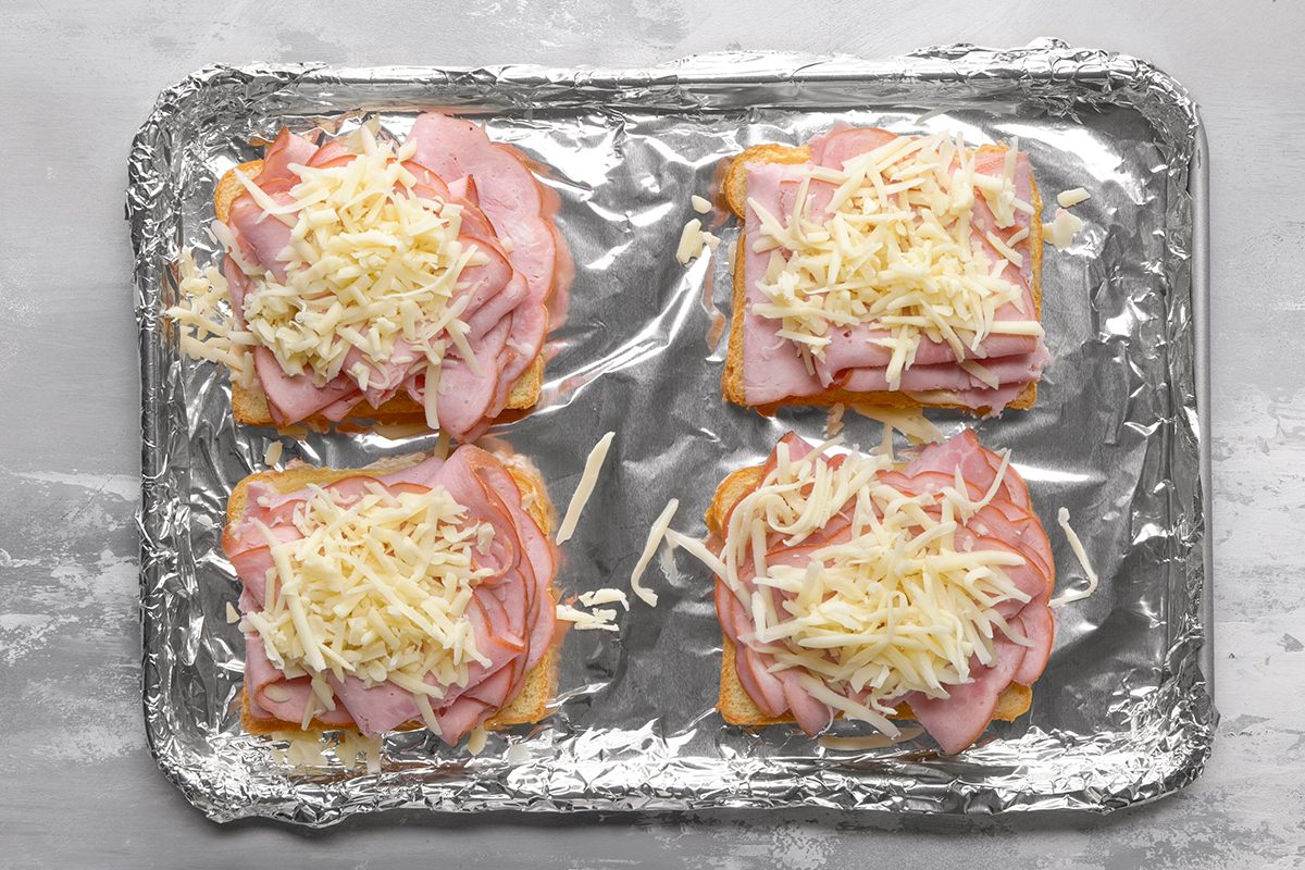 Four open-faced ham and cheese sandwiches on sliced bread are arranged on a foil-lined baking sheet, ready to be baked. Each sandwich is topped with sliced ham and shredded cheese.