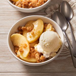 A white bowl filled with baked apple crisp topped with a scoop of vanilla ice cream sits on a light wooden surface next to two metal spoons and a checkered napkin.