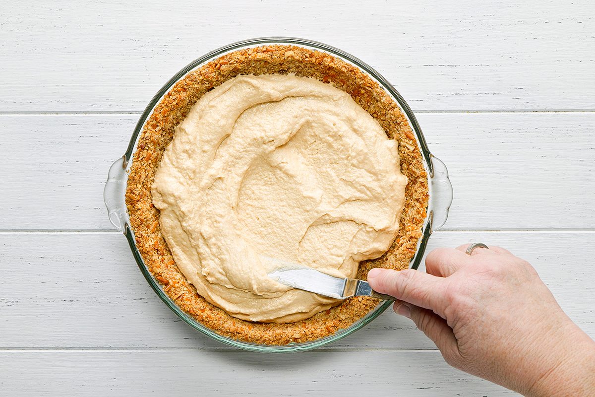 A hand spreads a creamy filling into a pie crust in a glass dish using a small spatula, on a white wooden surface.