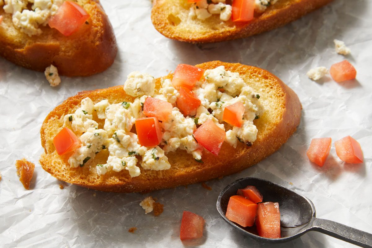closeup shot of a plate of Feta Bruschetta, nearby, there is a small black spoon containing additional diced tomatoes;