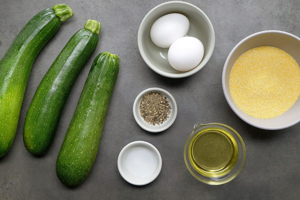 Three zucchinis, two eggs in a bowl, a bowl of cornmeal, small bowls of salt and pepper, and a cup of oil are arranged on a grey surface.