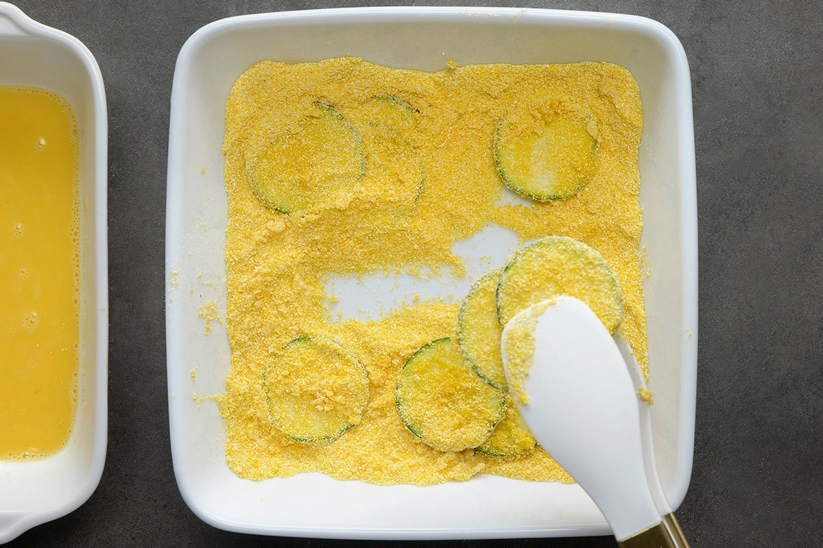 A white dish filled with yellow cornmeal and several slices of green zucchini being coated in the cornmeal, with a spatula partially covering some slices. Another dish with a yellow liquid is partially visible on the left.