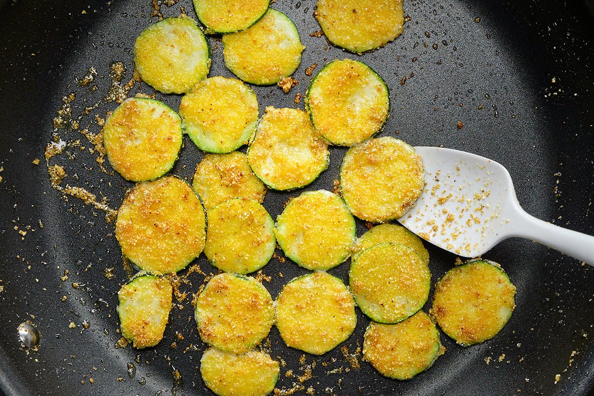 Sliced zucchini rounds coated in breadcrumbs and spices are being cooked in a skillet. A white spatula is seen stirring or flipping the zucchini pieces, which appear golden and crispy.