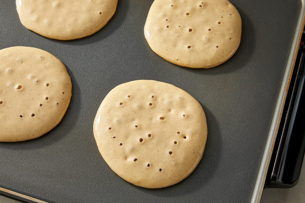 Four pancake batters are cooking on a griddle. The pancakes are light brown with bubbles forming on the surface, indicating they are in the process of being cooked. The griddle has a nonstick surface.