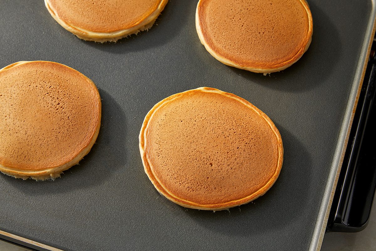 Four golden brown pancakes are cooking on a flat, nonstick griddle. The pancakes are evenly spaced and have smooth, round edges with lightly browned tops.