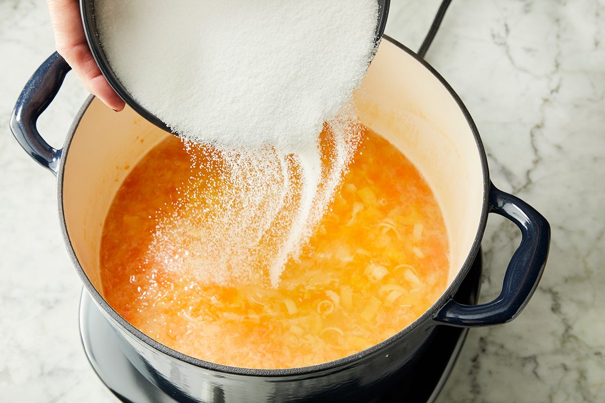 A hand pours a bowl of granulated sugar into a pot of orange-colored liquid simmering on a stove, likely preparing a sweet or savory dish.