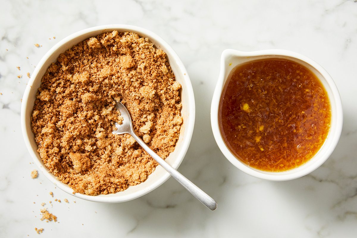 A bowl of crushed graham crackers with a spoon next to a small pitcher containing a melted butter and sugar mixture, both placed on a marble surface.