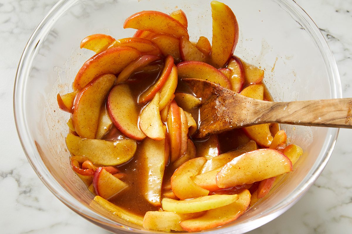Sliced apples coated in a cinnamon and sugar mixture are being stirred in a clear glass bowl with a wooden spoon, set on a white marble surface.