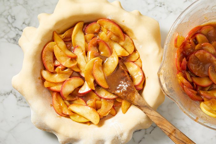 Sliced apples coated in spices are placed in an unbaked pie crust, with a wooden spoon resting inside. A mixing bowl with more apple slices is beside the pie on a marble countertop.