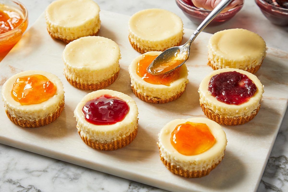 3/4 angle view shot of Jam-Topped Mini Cheesecakes; Cool for 10 minutes before removing from pan to a wire rack to cool completely; Refrigerate for at least 1 hour; Remove paper liners; top each cheesecake with 1 teaspoon jam; all arranged on a marble surface