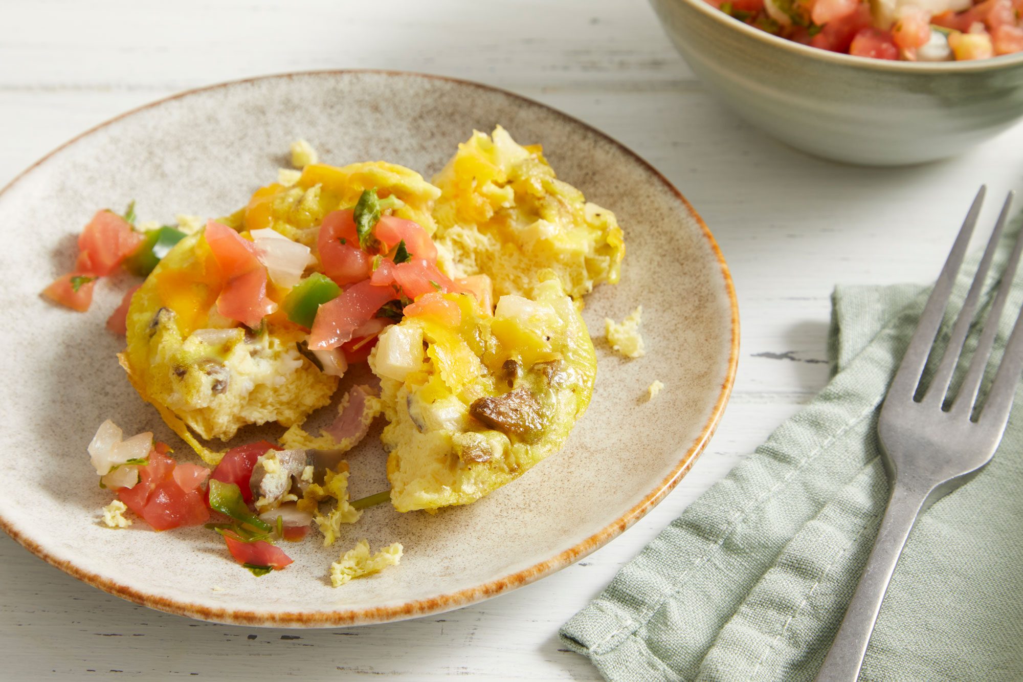 Close-up shot of Muffin-Tin Scrambled Eggs; served on a plate with pico de gallo; a fork rest on a napkin; all set on white wooden surface