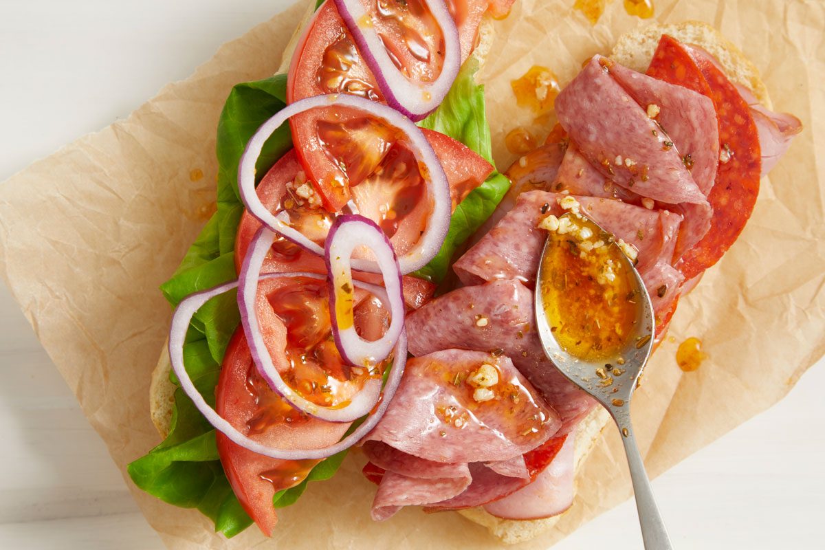 Close-up shot of Sub Oil and Vinegar Drizzle; being spooned over sandwiches; all set on a cream surface;