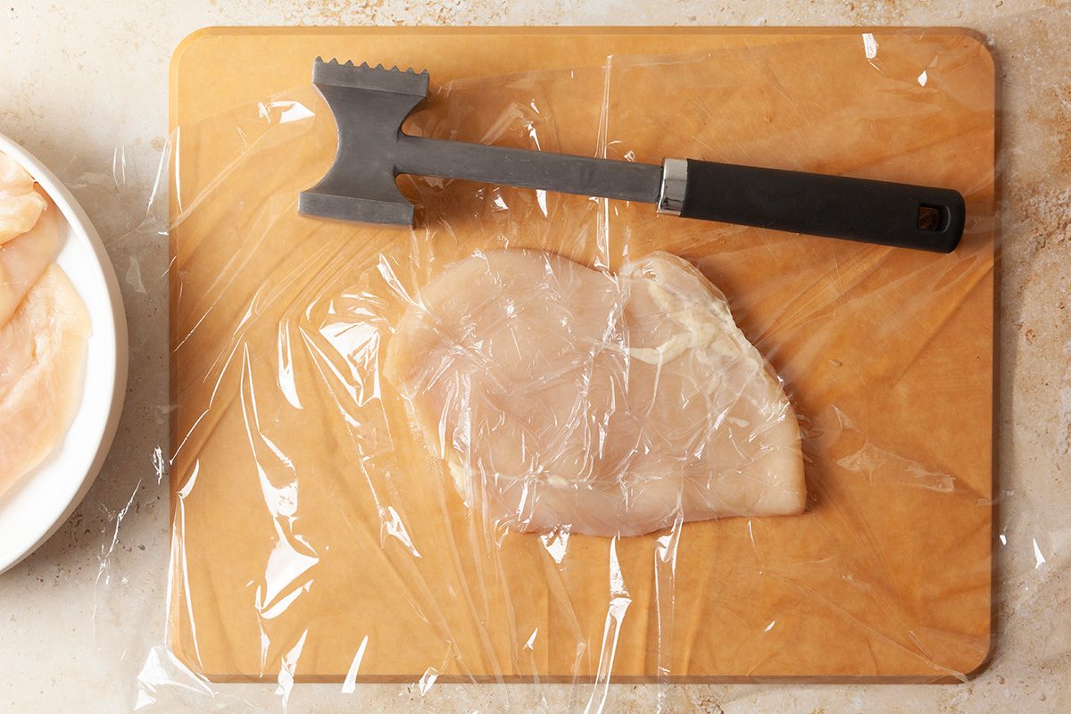 A raw chicken breast covered with plastic wrap on a wooden cutting board, next to a meat tenderizer.