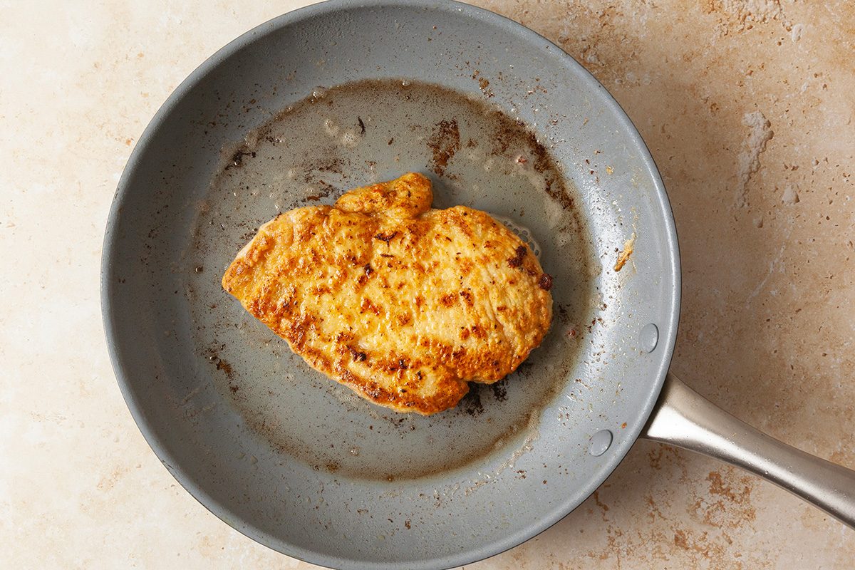 A cooked, seasoned chicken breast sits in a nonstick frying pan with some browned bits and oil residue on the surface. The pan rests on a light-colored countertop.