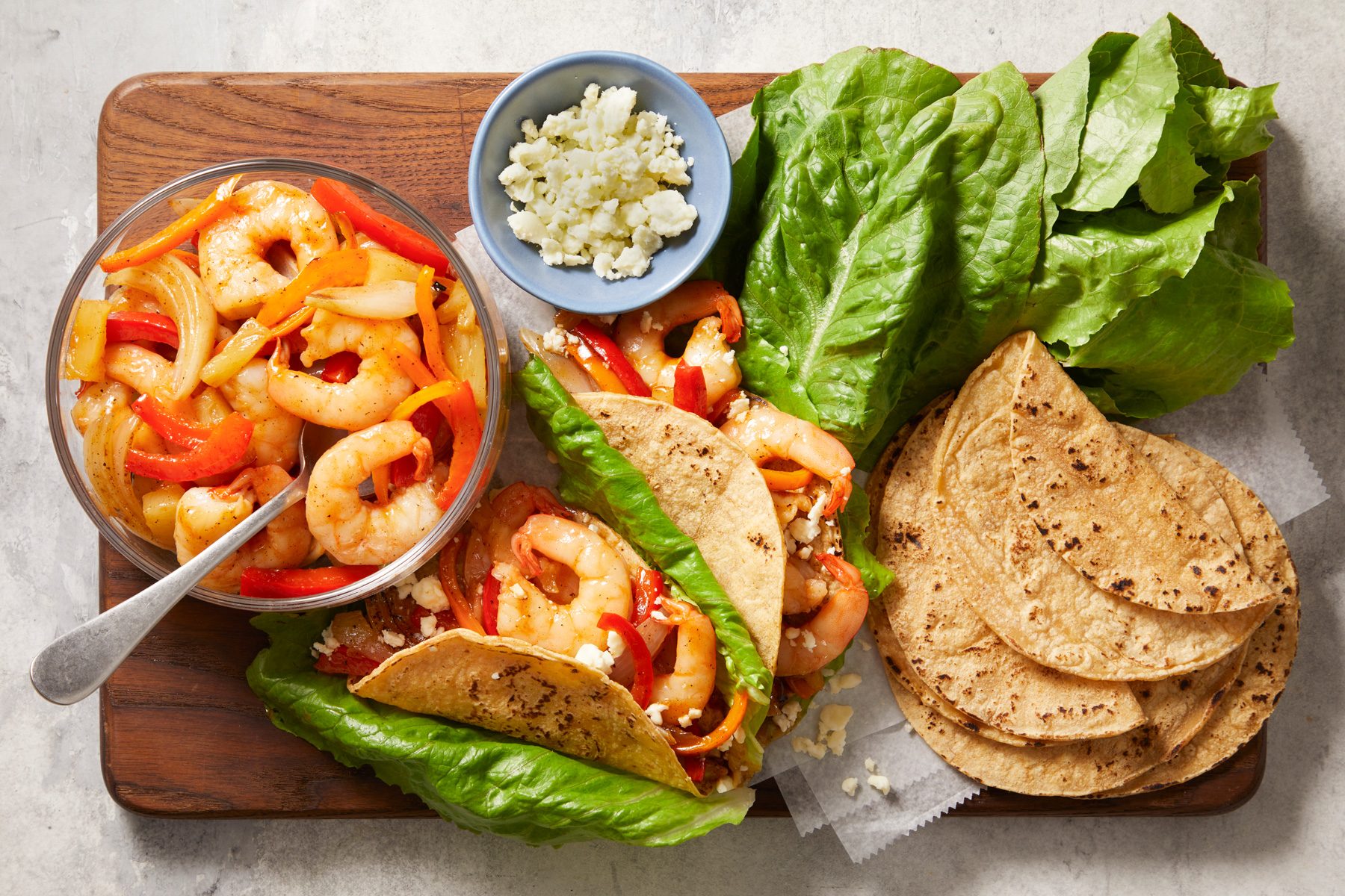 overhead shot of Pineapple Shrimp Tacos; On a wooden serving board, there is a transparent bowl filled with shrimp, bell peppers, and onions, Beside the bowl, several soft tortillas are neatly arranged with fresh lettuce leaves peeking out; A small blue bowl holds crumbled cheese