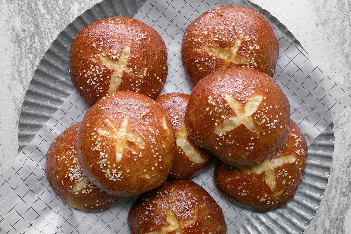 A group of golden-brown pretzel buns with a cross pattern on top and sprinkled with coarse salt, arranged on a round, textured metal tray with parchment paper underneath.
