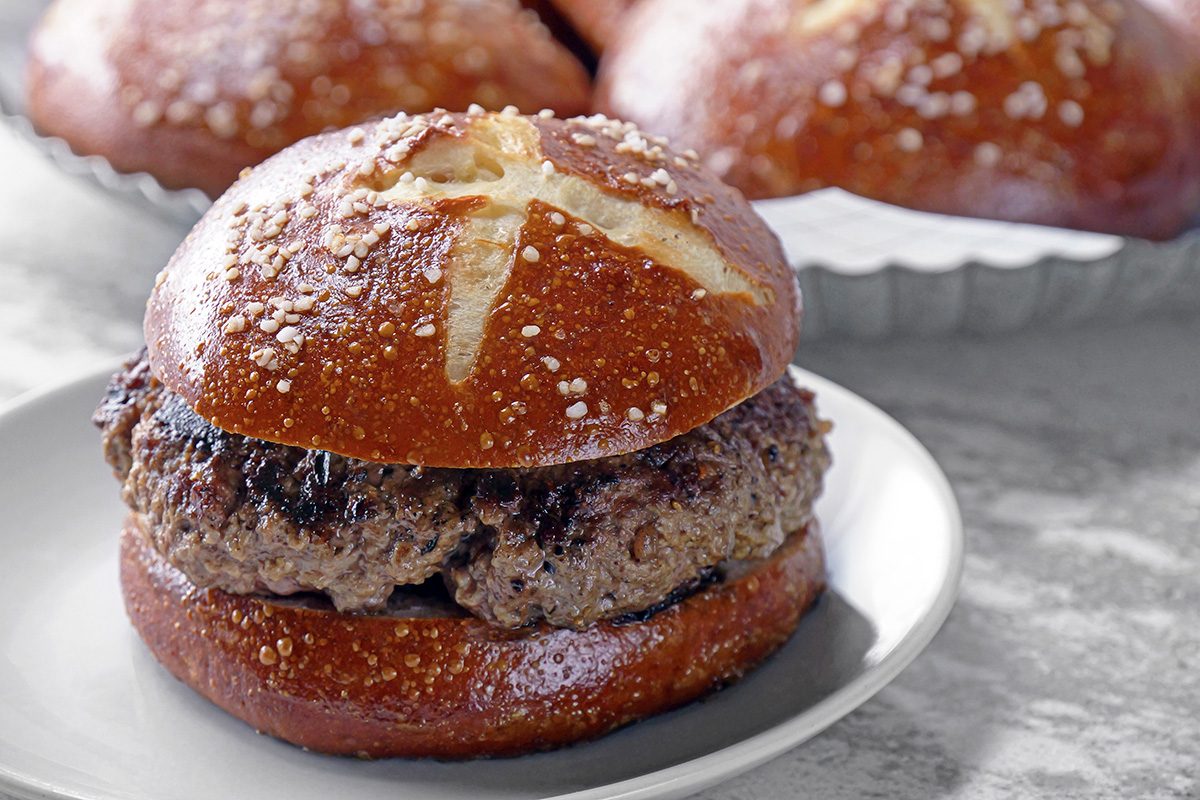 A juicy beef burger patty sits inside a pretzel bun, topped with coarse salt and a cross cut into the top. The burger is served on a white plate with more pretzel buns in the background.