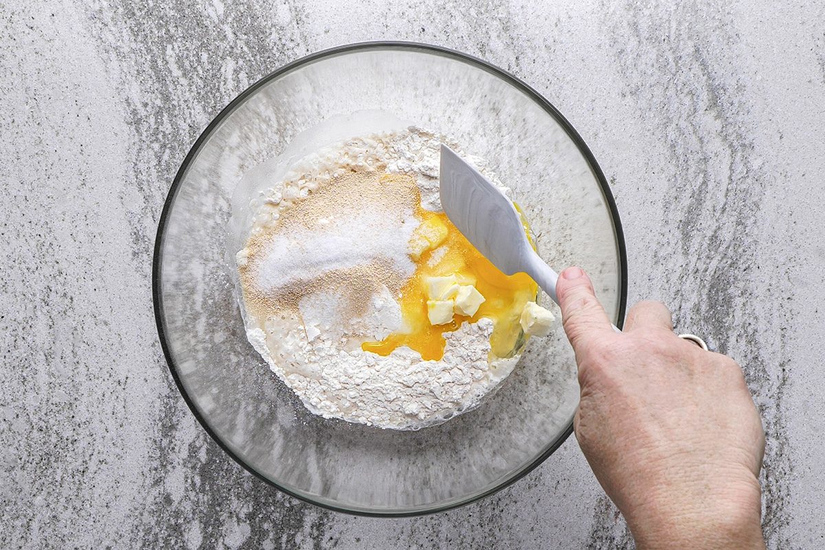 A hand holding a spatula mixes flour, sugar, egg, and butter in a glass bowl on a gray countertop.