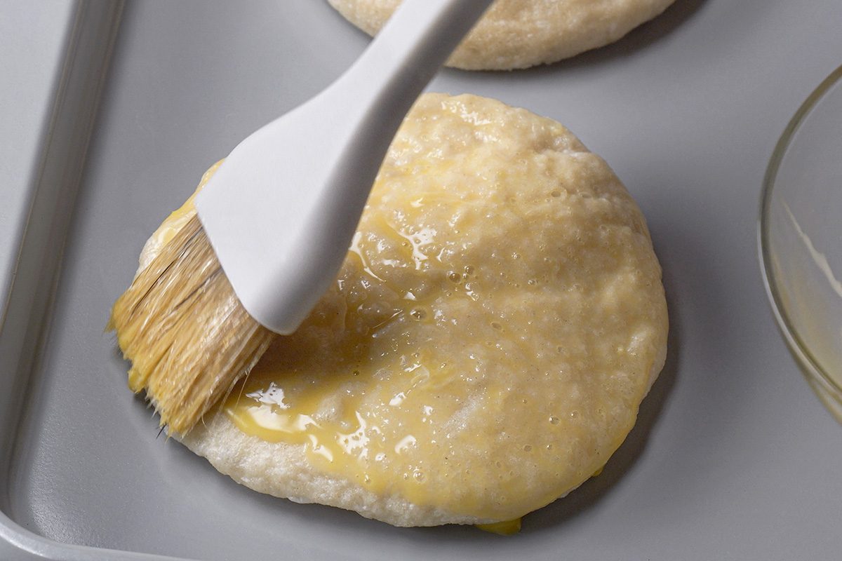 A pastry brush is spreading melted butter onto a round, uncooked biscuit dough on a baking sheet.