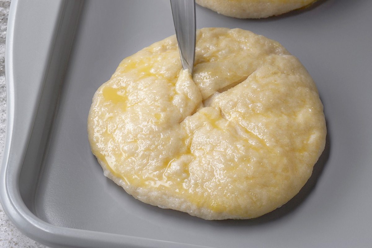 A knife scoring a round, unbaked, egg-washed dough on a gray baking tray, preparing it for baking.