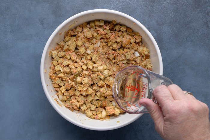 overhead shot of A white bowl containing a mixture of light brown bread cubes, small pieces of light pink meat, and translucent white pieces of onion or celery; a hand is pouring a clear liquid, possibly broth or water, from a clear glass measuring cup into the bowl; the bowl is positioned over a solid, muted blue gray surface
