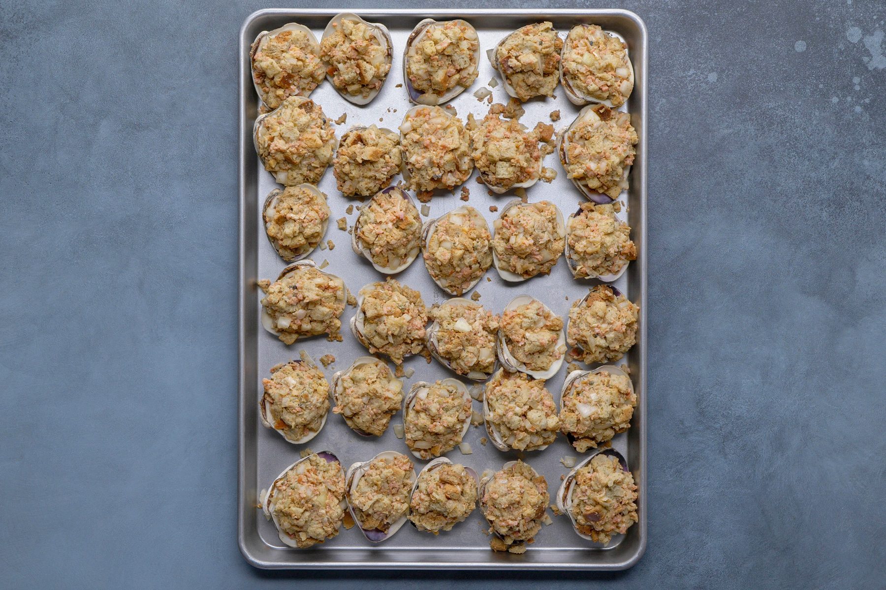 overhead shot of a silver baking sheet filled with numerous stuffed clam shells, arranged in neat rows; each clam shell is filled with a light brown, breadcrumb based stuffing, likely containing chopped seafood and vegetables; the baking sheet has a slightly raised rim and rests on a solid, muted blue gray surface