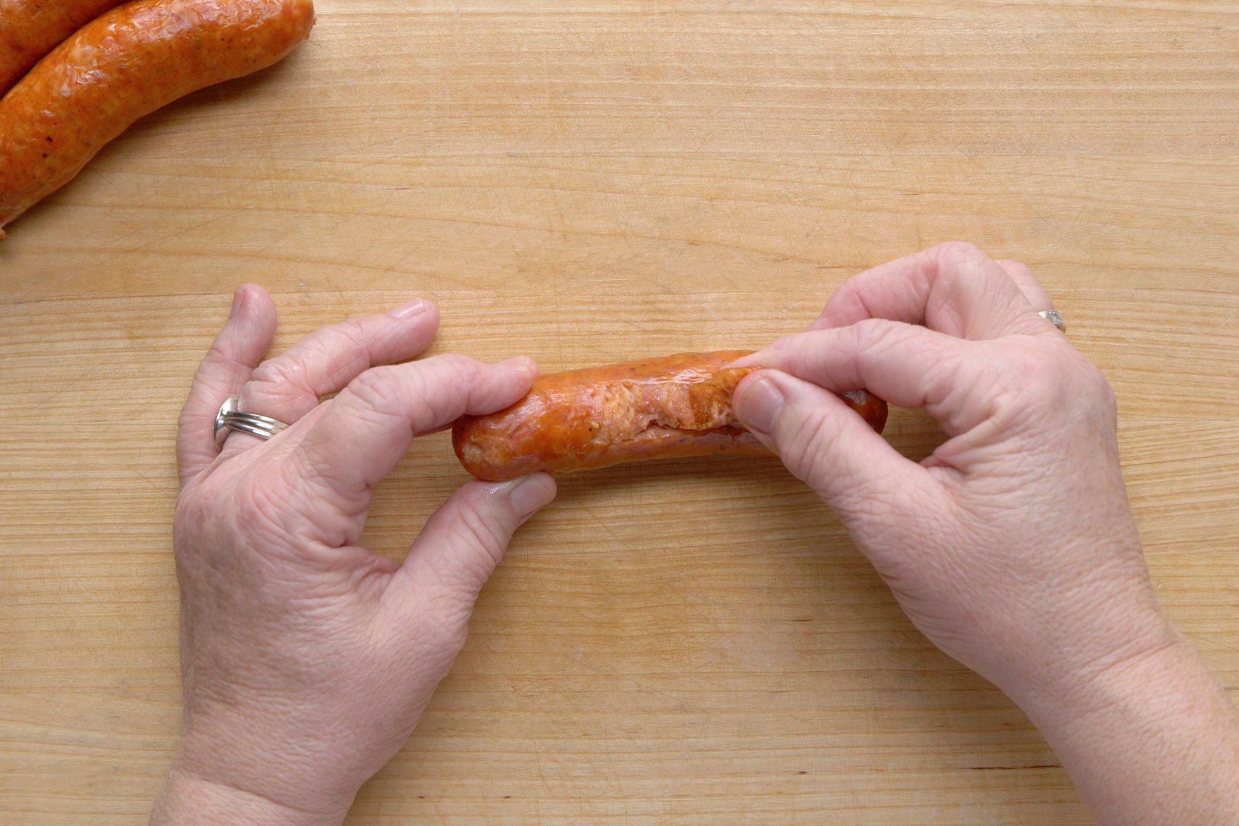 overhead shot of Two hands are shown manipulating a cooked sausage over a light brown wooden surface; the hands are gently squeezing or twisting the sausage in the middle; two additional sausages are visible in the upper left corner of the frame, lying on the same wooden surface