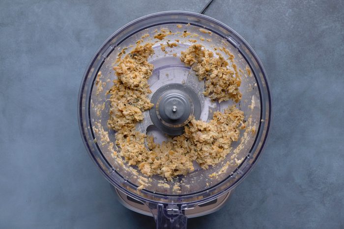 overhead shot of the inside of a clear plastic food processor bowl containing a chunky, light brown mixture, possibly homemade peanut butter or a similar nut butter; the bowl is sitting on the base of the food processor, which is partially visible at the bottom of the frame, and the background is a solid, muted blue gray surface