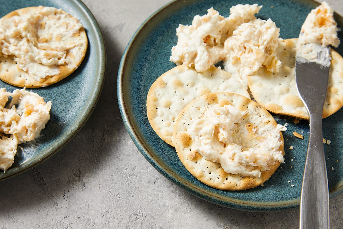 A blue plate with round crackers topped with a creamy spread, and a knife with more spread resting on the plate. Another plate with crackers and spread is partially visible beside it.