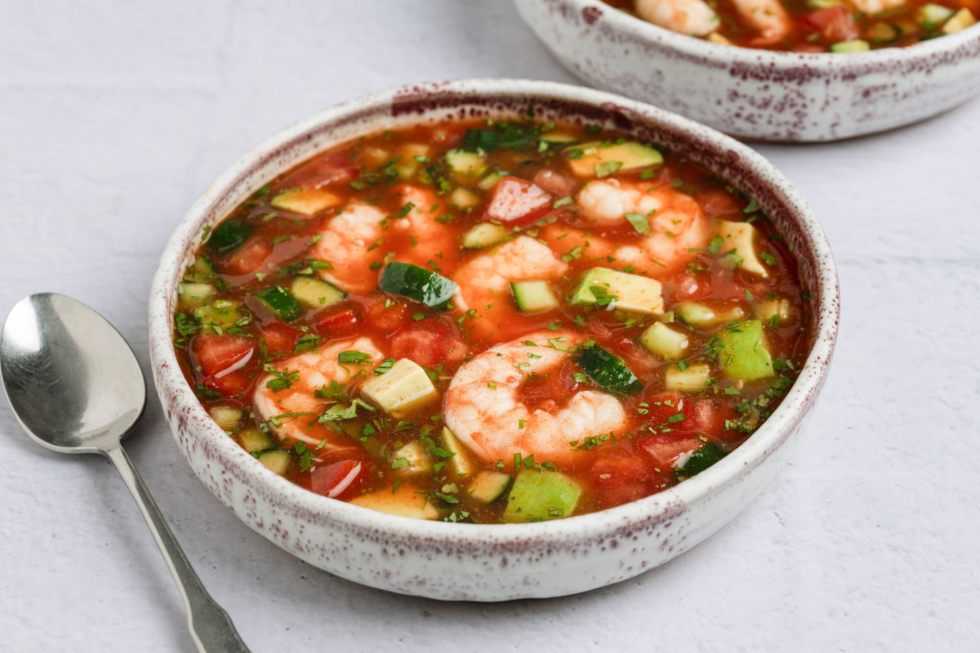 overhead shot of a dish called Shrimp Gazpacho; in two shallow bowls, next to the bowls are two shiny silver spoon, the background is a soft, light gray