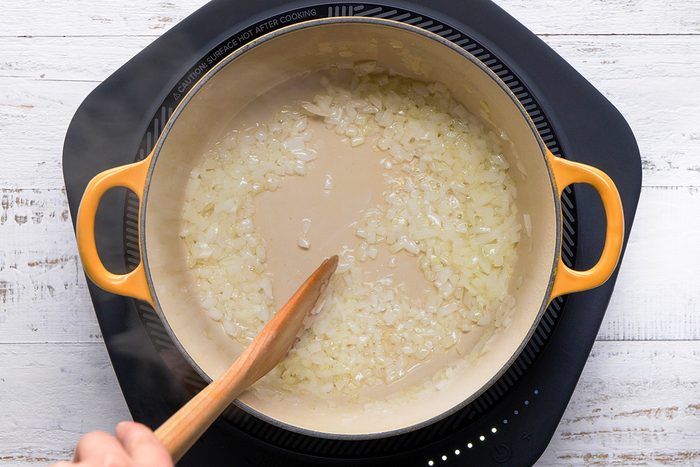A cook using a wooden spoon to stir onions in a pot, with a blurred kitchen background and steam visible.