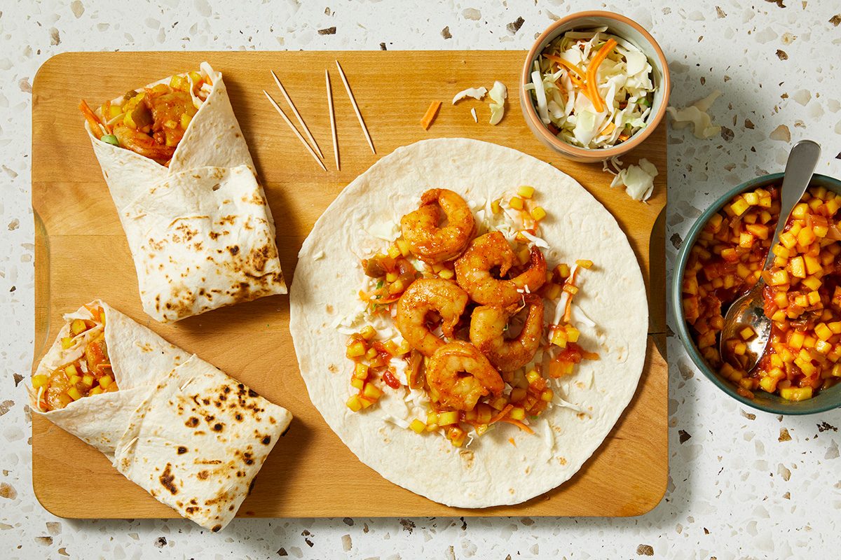 A cutting board with a shrimp wrap, two halved shrimp burritos, shredded cabbage in a bowl, and a bowl of pineapple salsa on a light speckled surface.