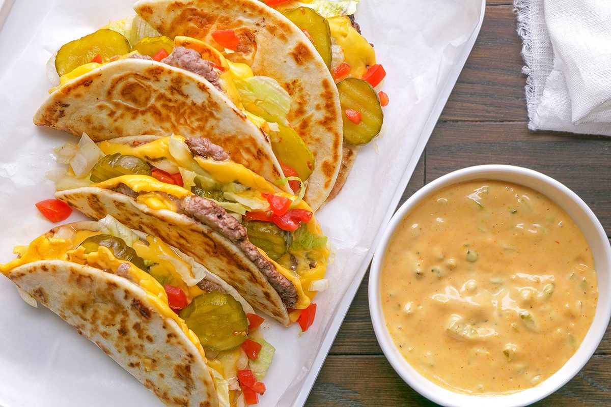 Three cheesy ground beef tacos with pickles, diced tomatoes, and onions are arranged on a white plate next to a bowl of creamy cheese dip on a wooden table.