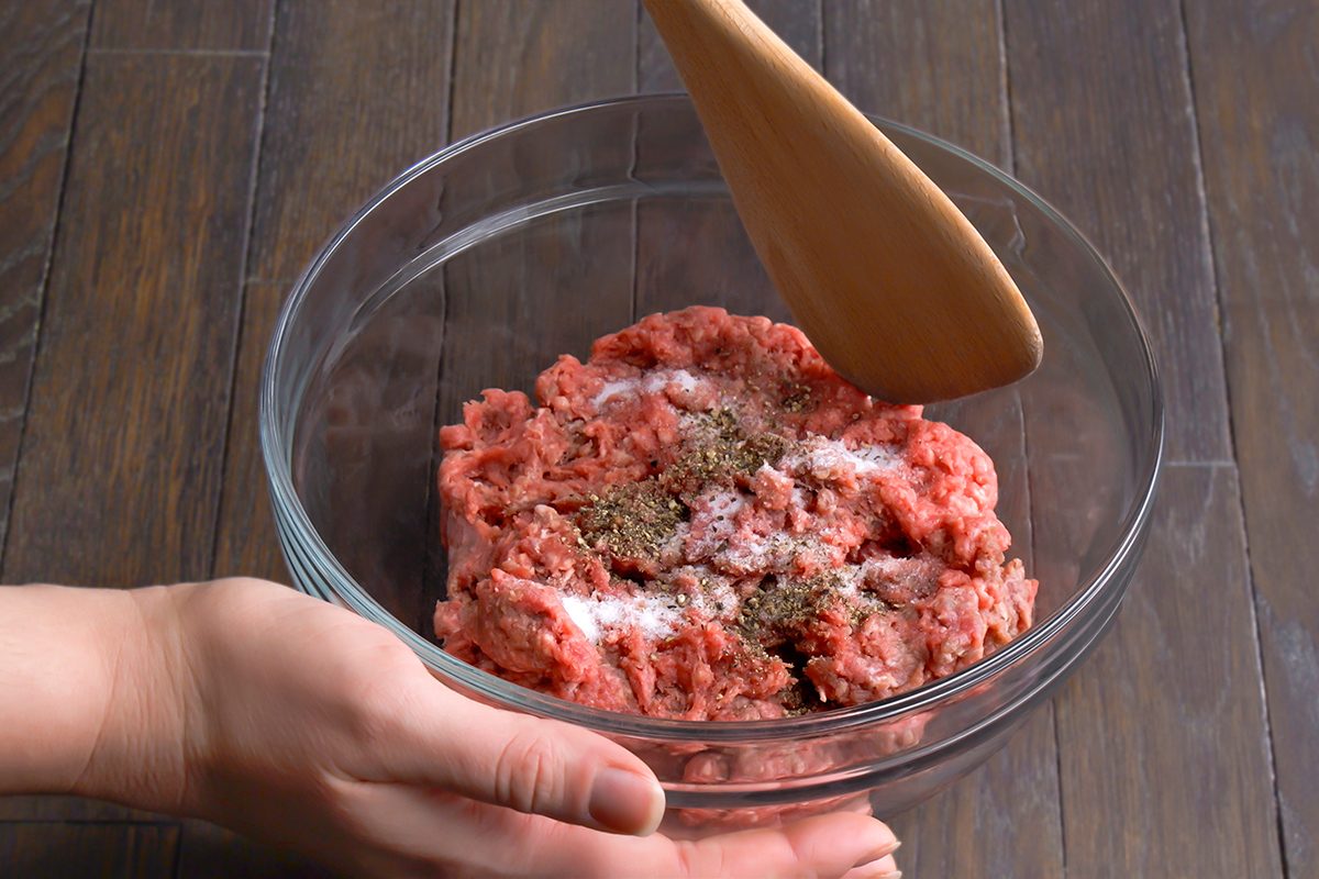 A hand holds a glass bowl filled with ground meat, salt, and pepper, while a wooden spatula is poised above the mixture. The bowl is on a dark wooden surface.