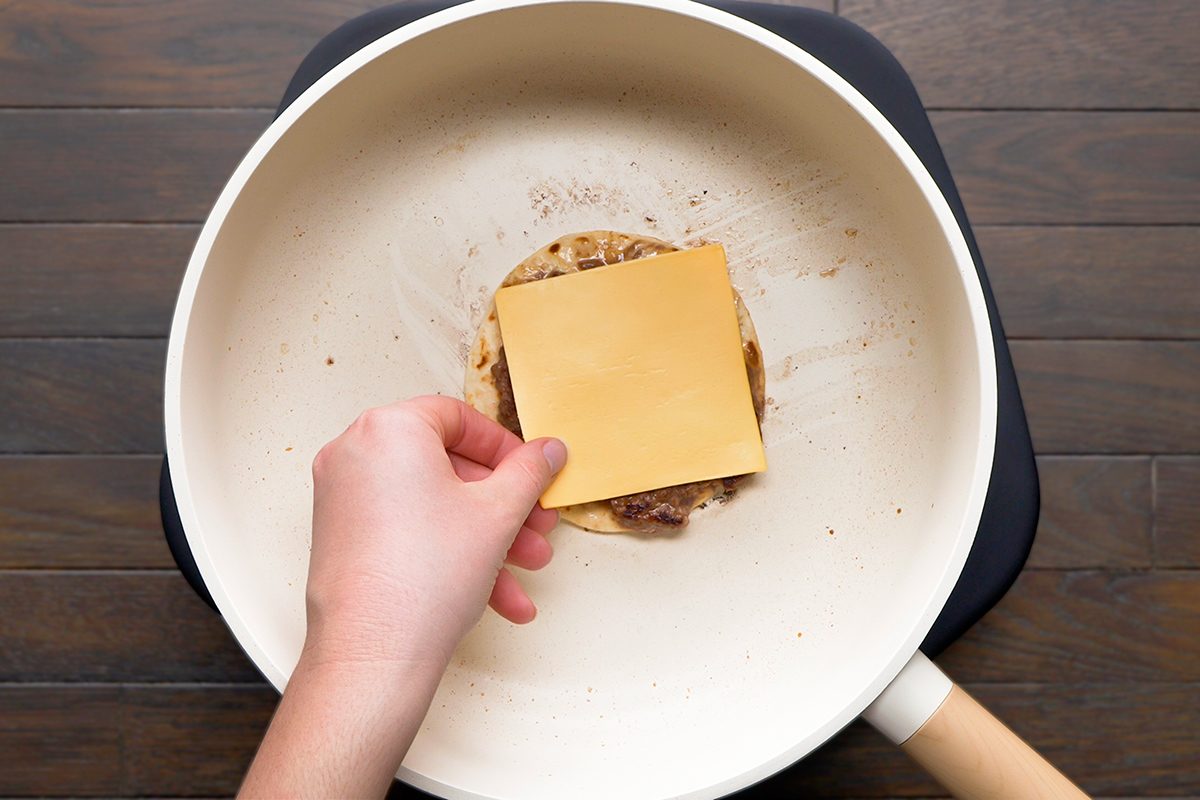A hand places a slice of cheddar cheese on a burger patty cooking in a light-colored frying pan on a dark stovetop.