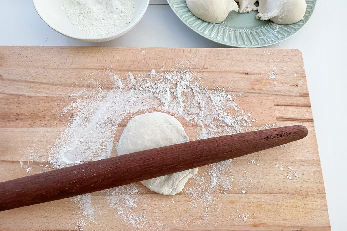 A wooden rolling pin flattening a small piece of dough on a floured wooden surface, with extra dough and a bowl of flour in the background.